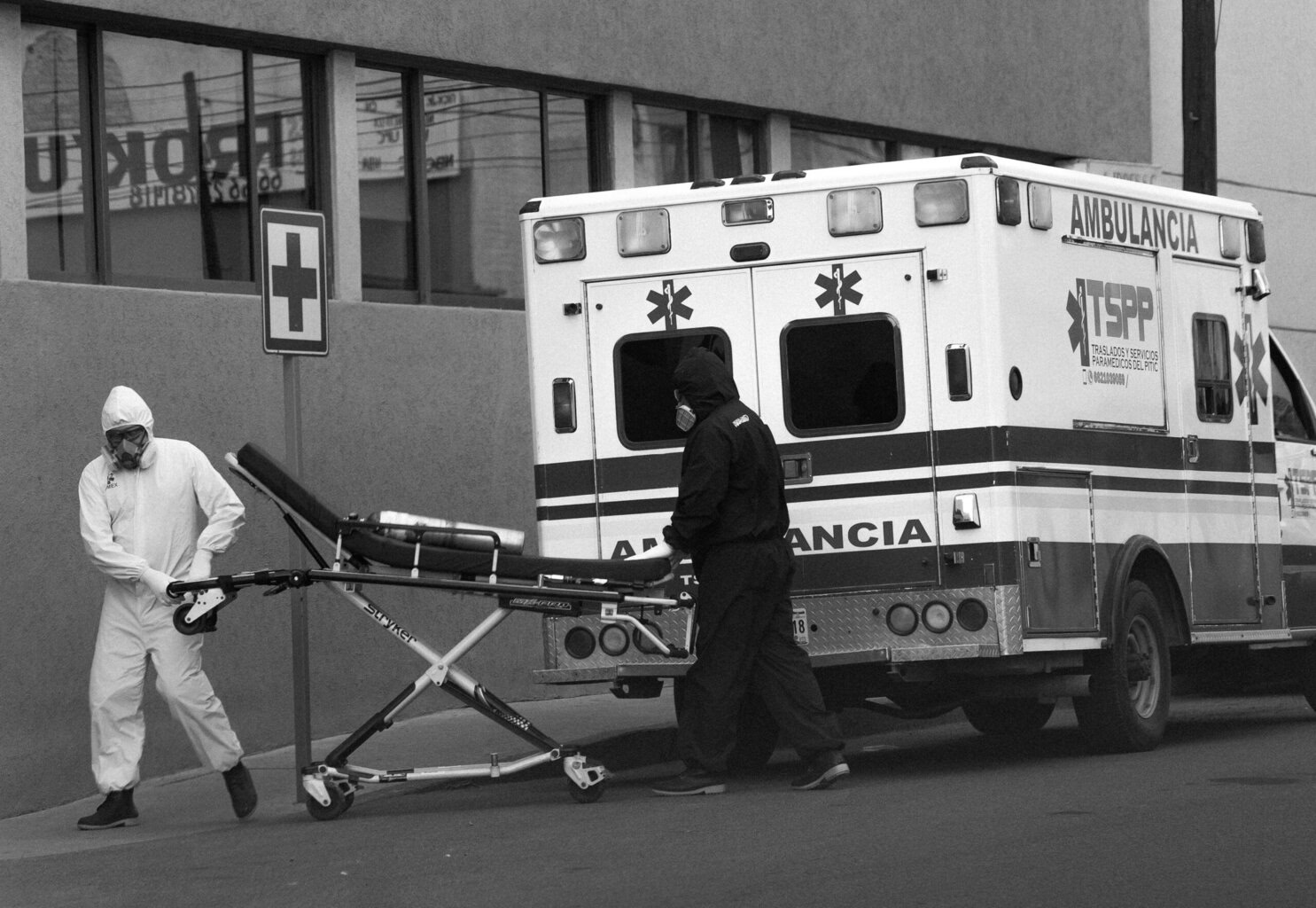Dos personas con trajes de protección llevan una camilla vacía hacia una ambulancia aparcada frente a un edificio.