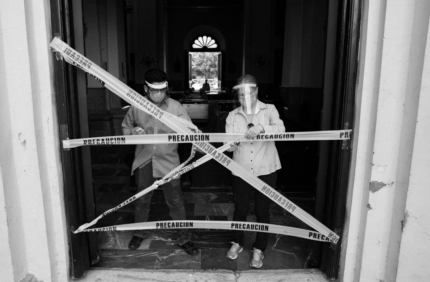 Dos personas que llevan protectores faciales se encuentran detrás de una cinta de precaución cruzada con la inscripción «PRECAUCIÓN» en la entrada de un edificio.