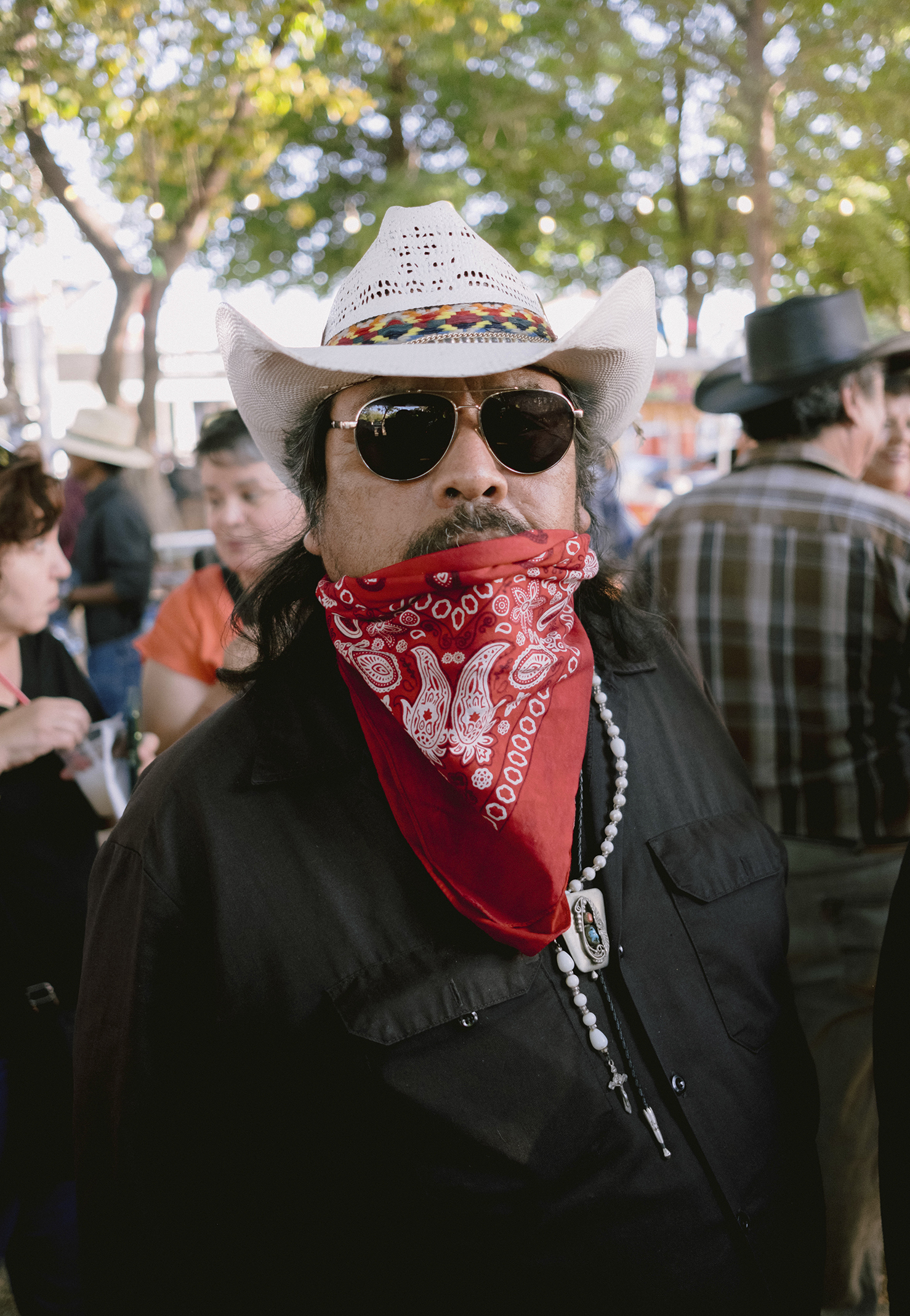 Man in a black shirt, white cowboy hat, dark sunglasses, red bandana, and necklace with large pendant standing outdoors among other people with trees in the background.