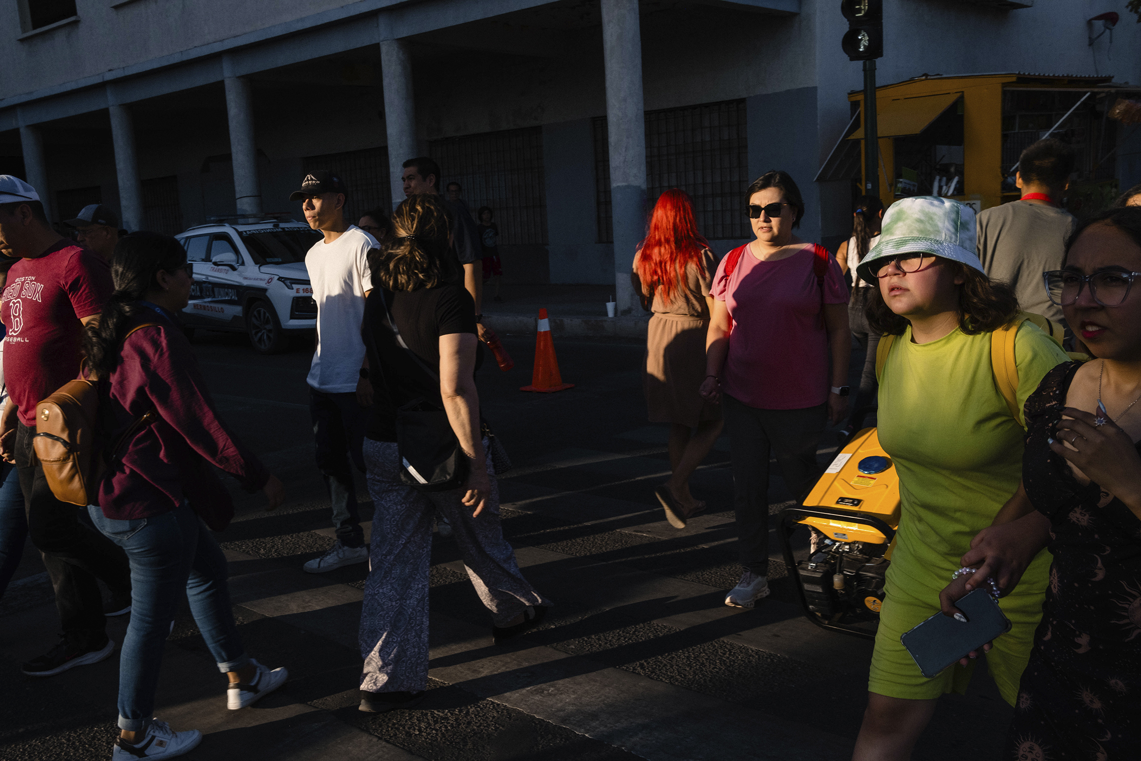 People wearing various outfits walk in different directions on a street with sunlight casting shadows. A yellow robot-like object is present beside one of the pedestrians.