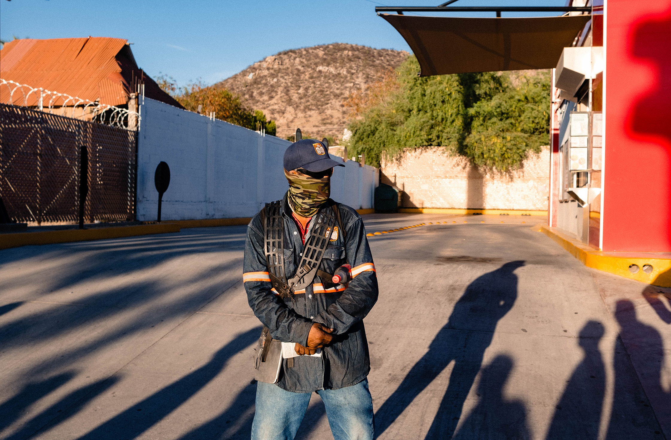 A person wearing a mask and uniform stands with clasped hands in a sunlit area near a gated entrance, with a hilly background and shadows cast on the ground.