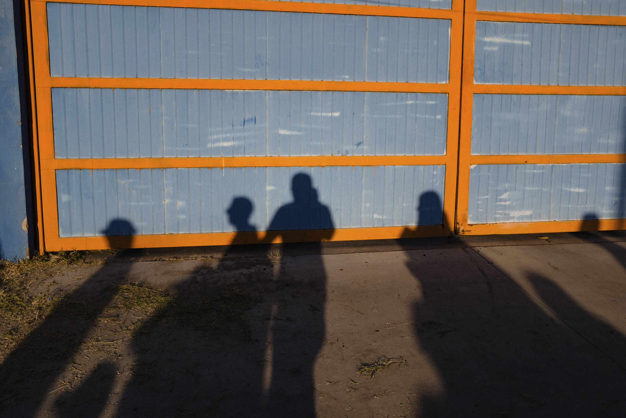 Shadows of several people are cast on the ground and a large blue and orange gate behind them, under bright sunlight.