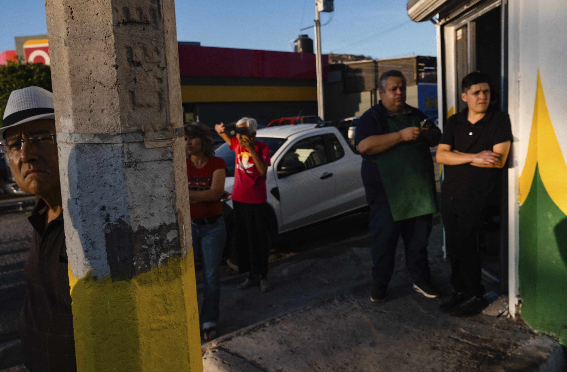 People are standing outside near a building and a white truck, some in conversation, and one person shielding their eyes from the sun. A pole with yellow paint in the foreground partially obscures the view.