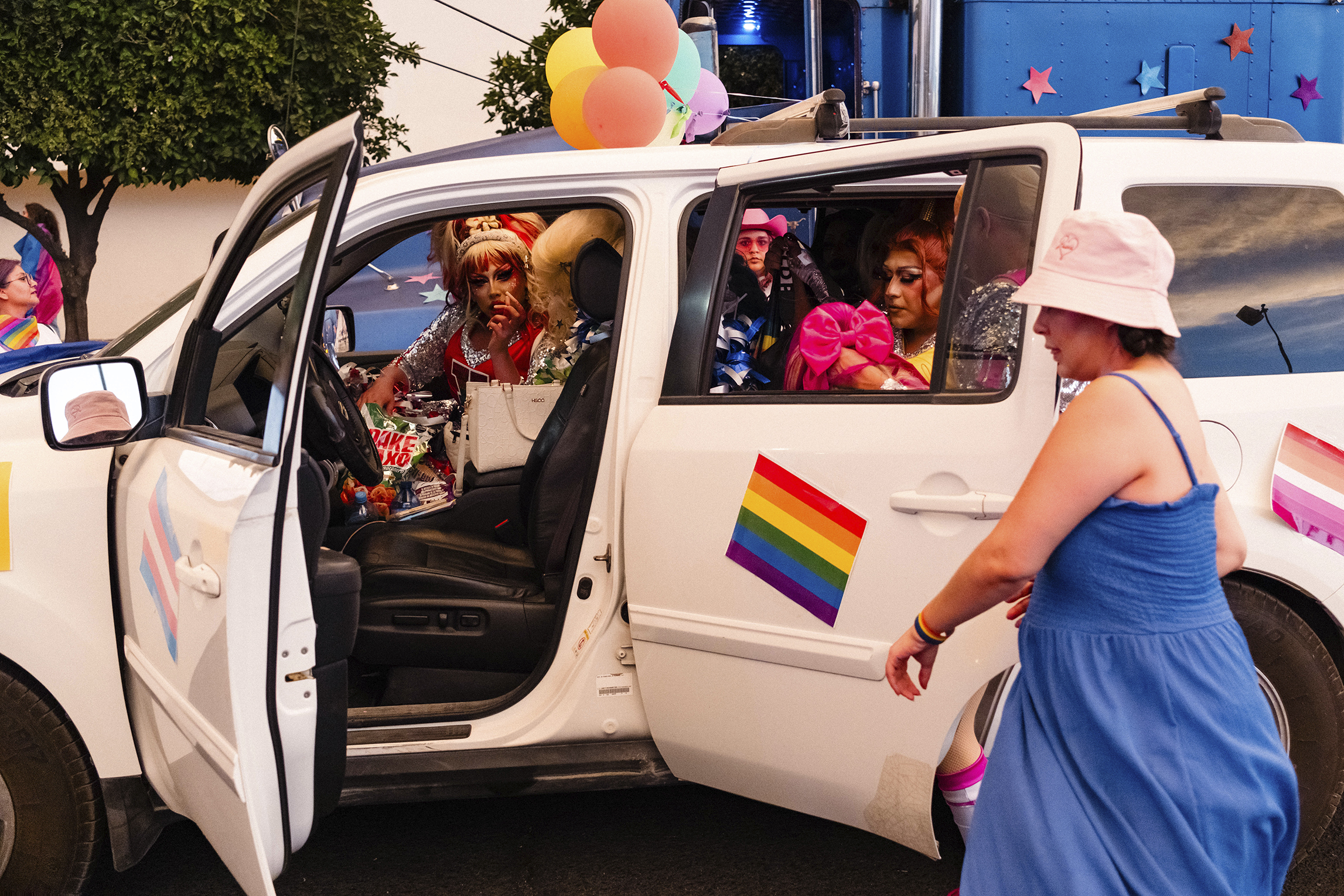 A person in a blue dress walks past a decorated white vehicle with pride flags and balloons. Several individuals in colorful outfits are inside the vehicle.