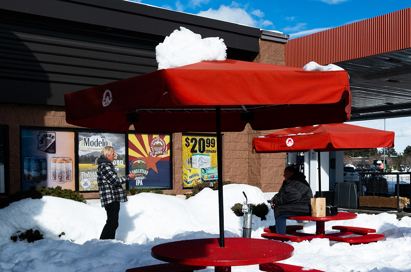 Two people are by red outdoor tables with snow on top, next to a building displaying posters advertising beer; the ground and tables are covered with snow.