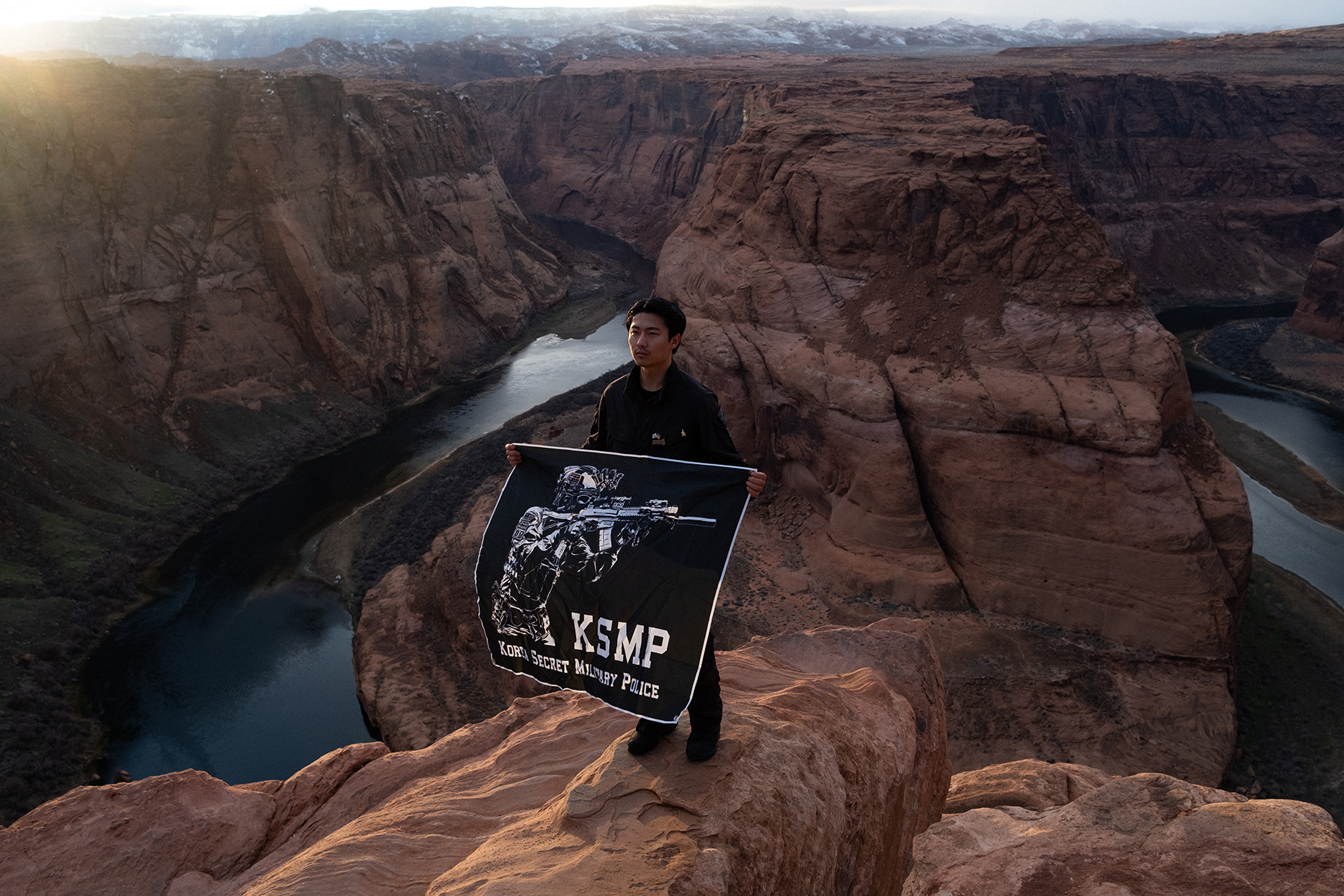 A person stands on a cliff holding a banner with text and an image of a sniper, against the backdrop of a deep canyon and river below.