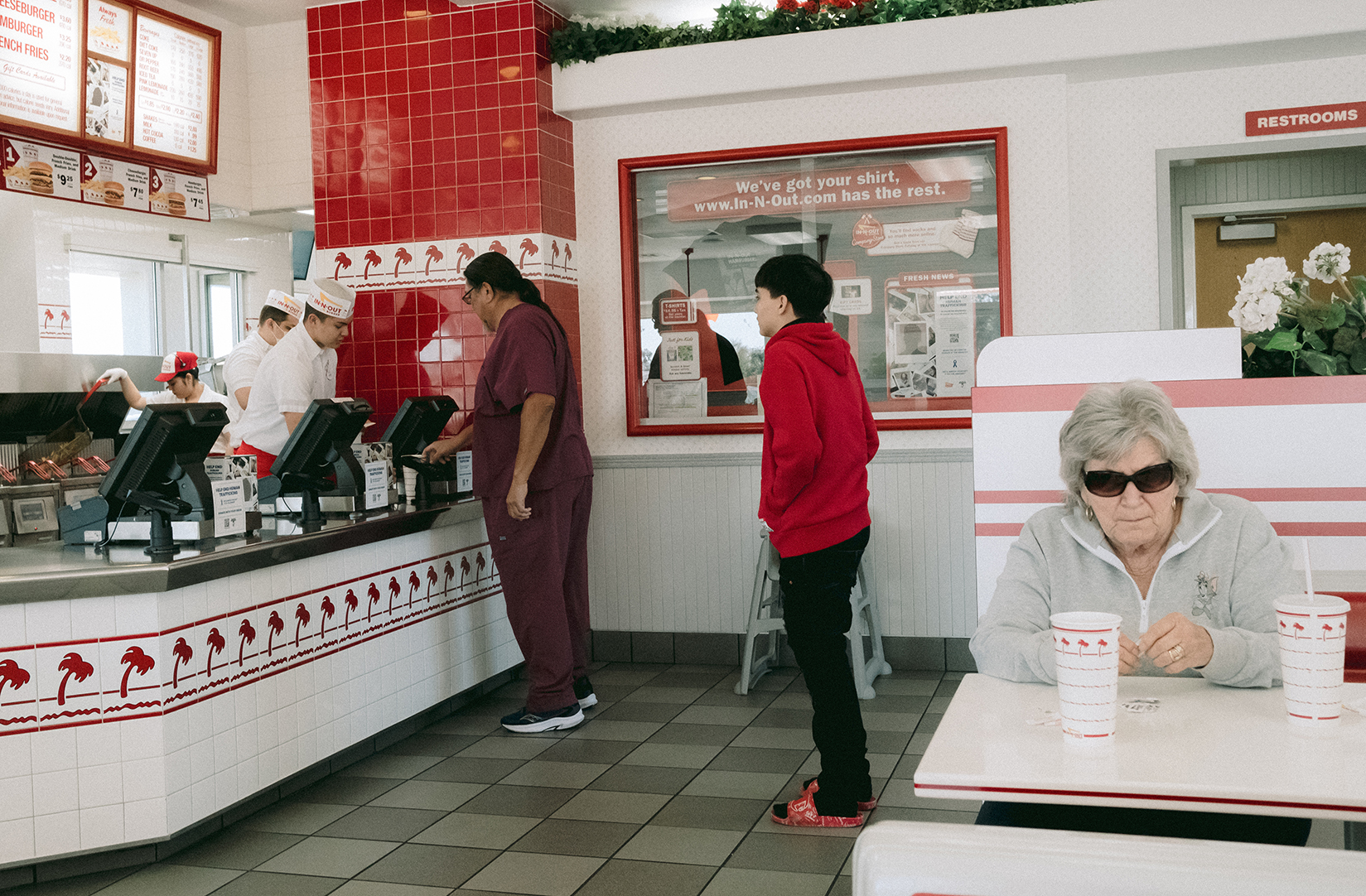 People standing and ordering food at the counter in a fast-food restaurant, with a woman sitting at a booth in the foreground.