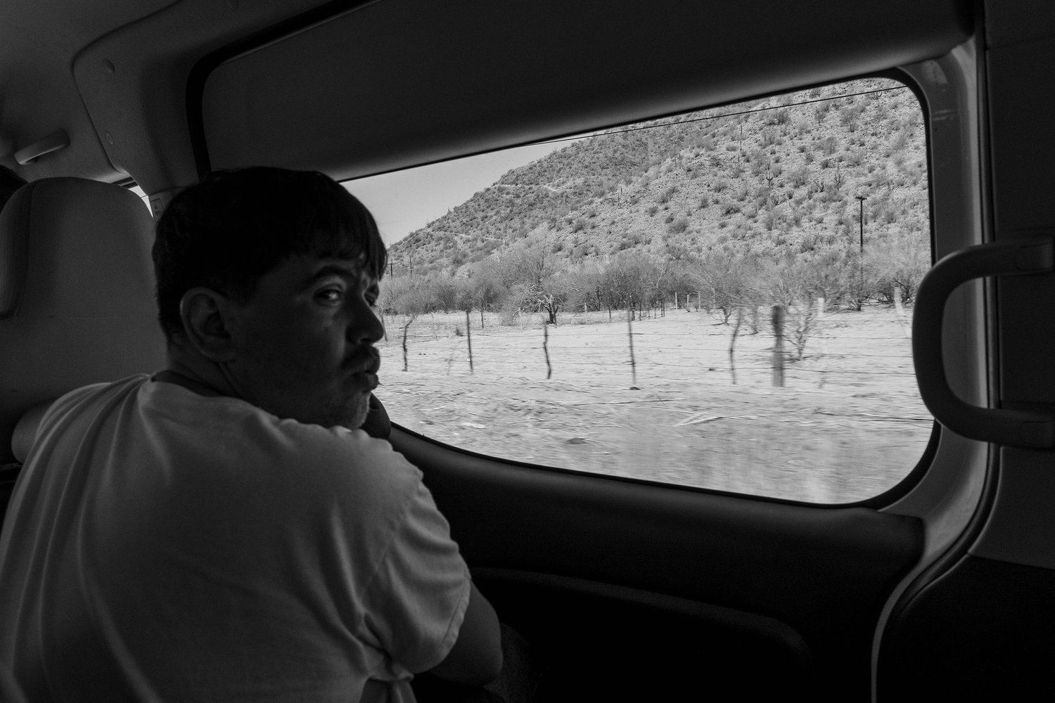 A person looks out through a vehicle window at a desert landscape with sparse trees and hills. The image is in black and white.