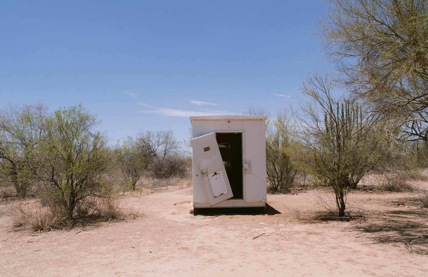 A small, white shed with an open door stands in a dry, sparse desert landscape under a clear blue sky.