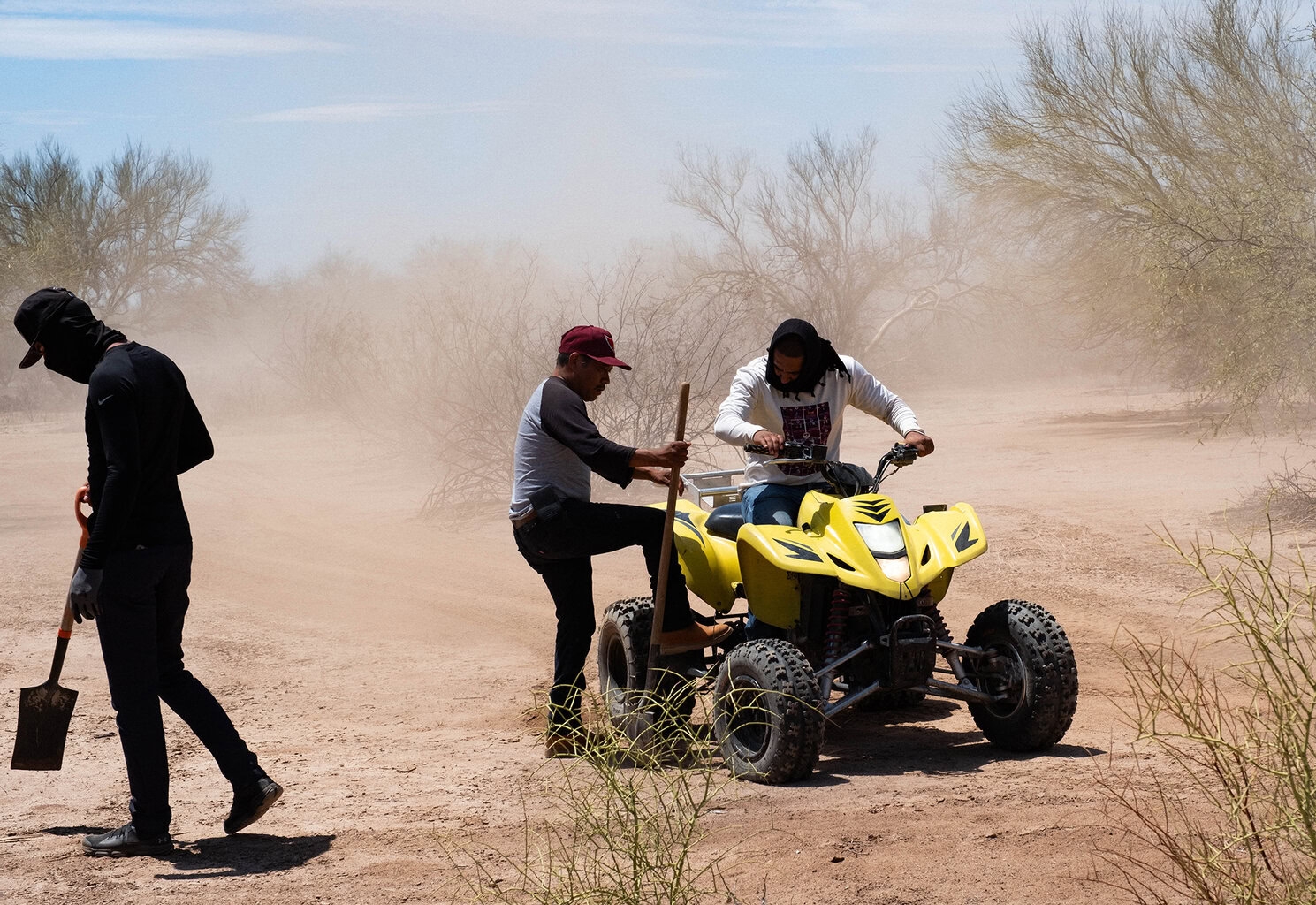 Three people in a dusty desert setting, one riding a yellow ATV, another assisting, and a third walking while holding a shovel. Sparse vegetation is visible in the background.