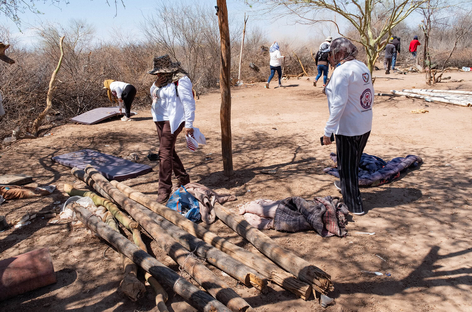 A group of people wearing protective clothing search a dry, desert-like area with scattered blankets and wooden poles.