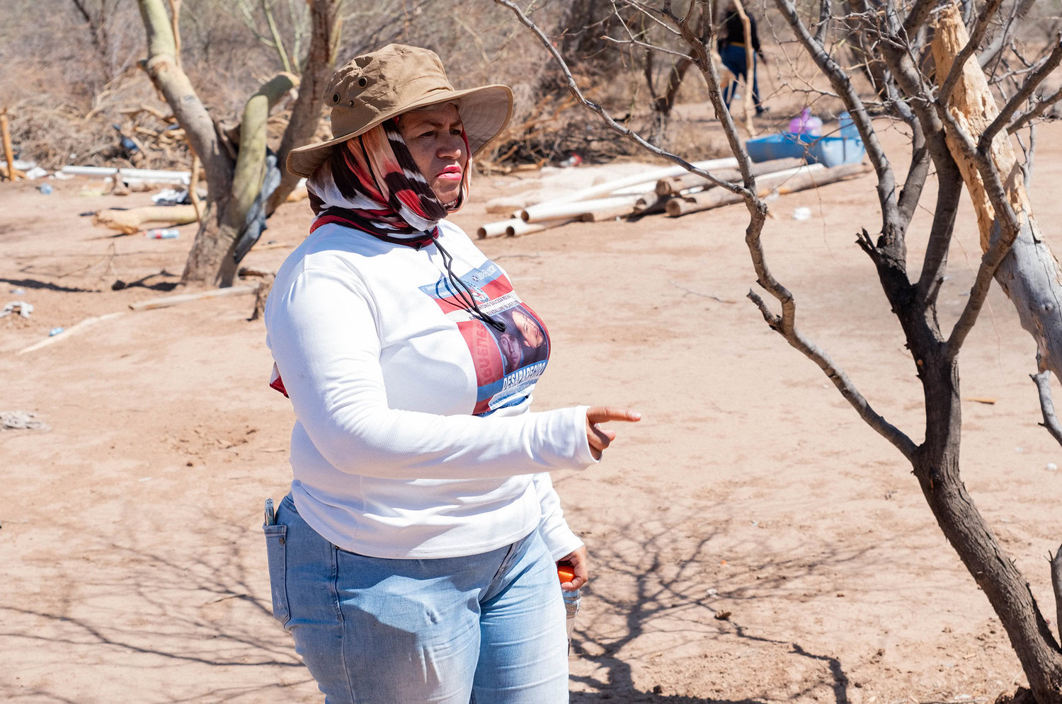 Person in a sun hat and scarf points at a dry, barren landscape with sparse trees and debris scattered around.