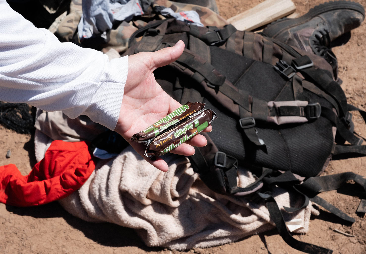 Hand holding two candy bars in front of a backpack and clothing on the ground.