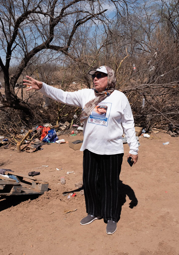 Person in a white shirt and cap gestures outdoors in a dry, wooded area with scattered debris around them.