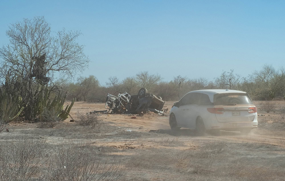 White car driving towards an overturned vehicle in a dry, barren landscape with sparse vegetation.