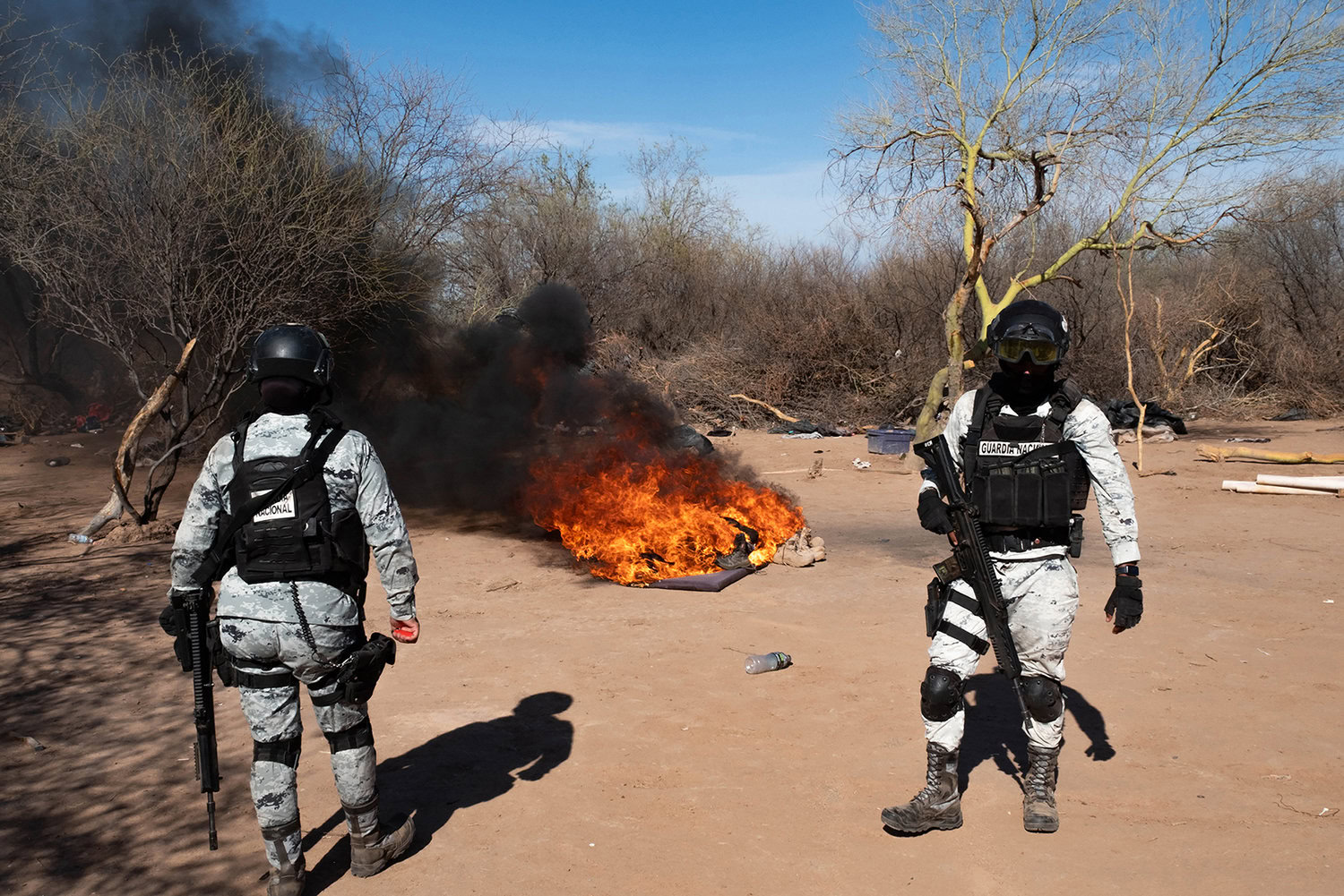 Two armed uniformed individuals stand in a desert landscape near a fire, with smoke rising into the sky. Sparse vegetation is seen in the background.