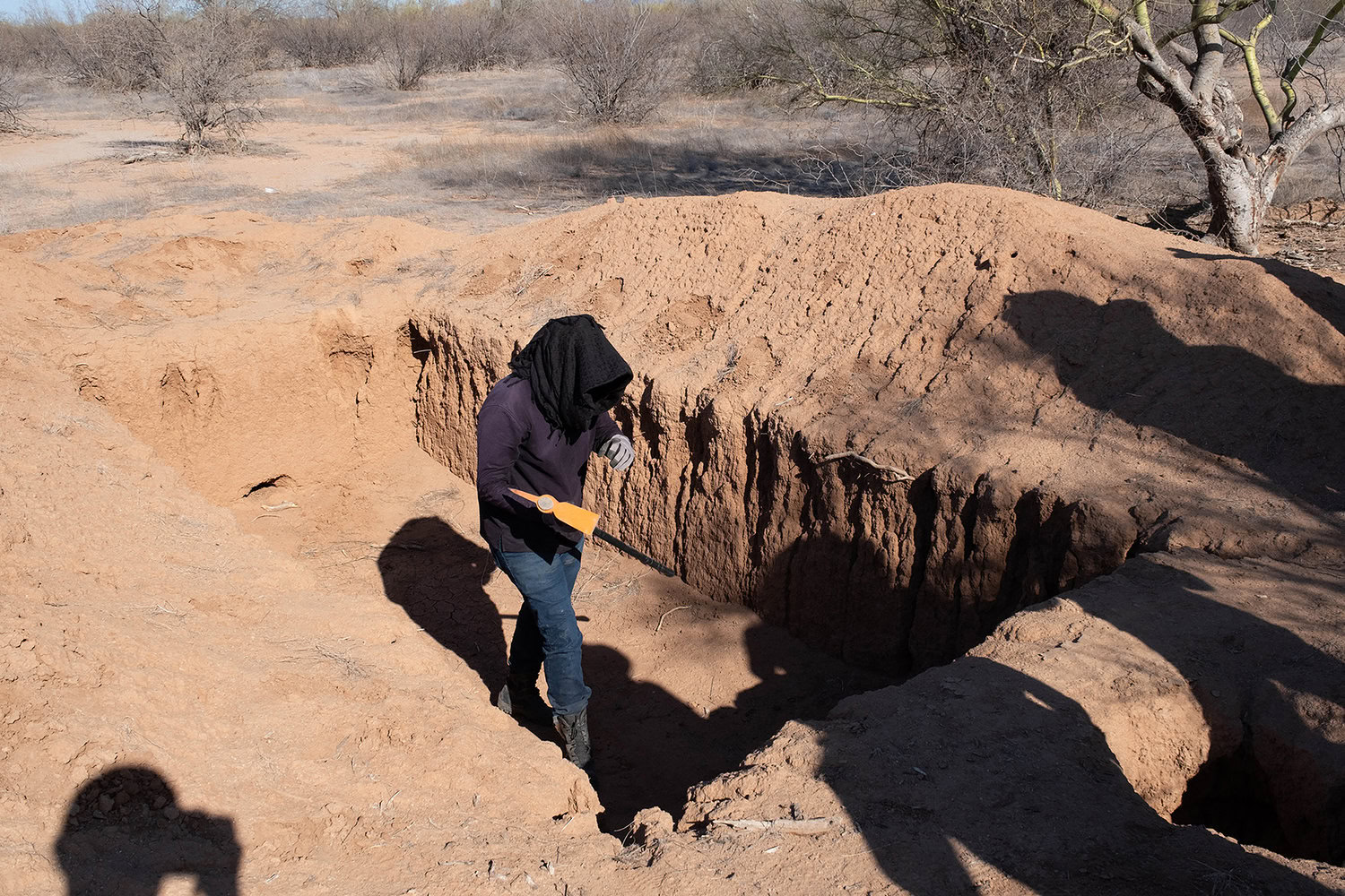 A person stands in a deep, excavated pit in a desert landscape, using a tool. Shadow of another person is visible in the foreground. Sparse vegetation surrounds the area.