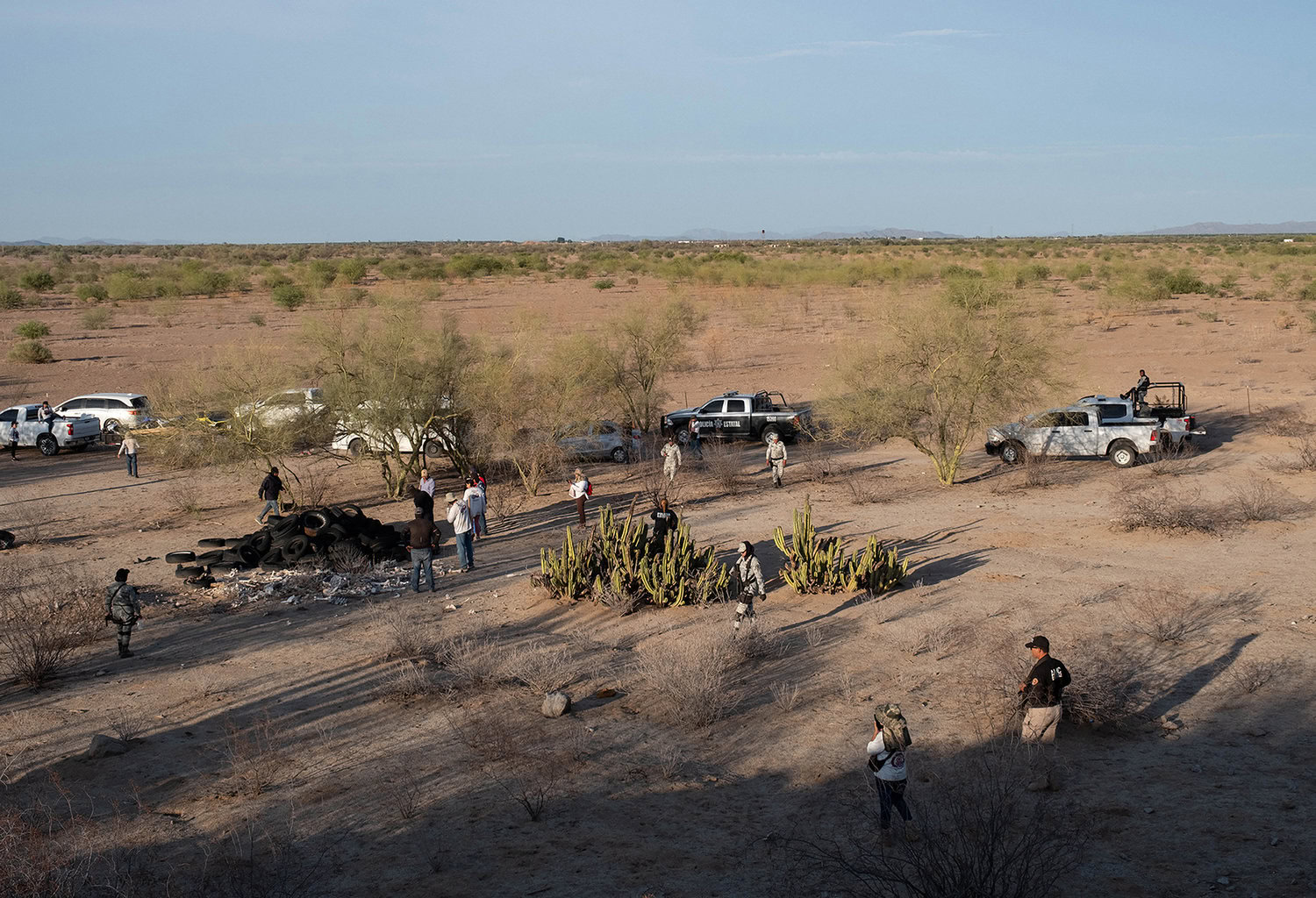 A group of people and vehicles are scattered in a desert landscape with cacti, tire piles, and sparse vegetation under a blue sky.