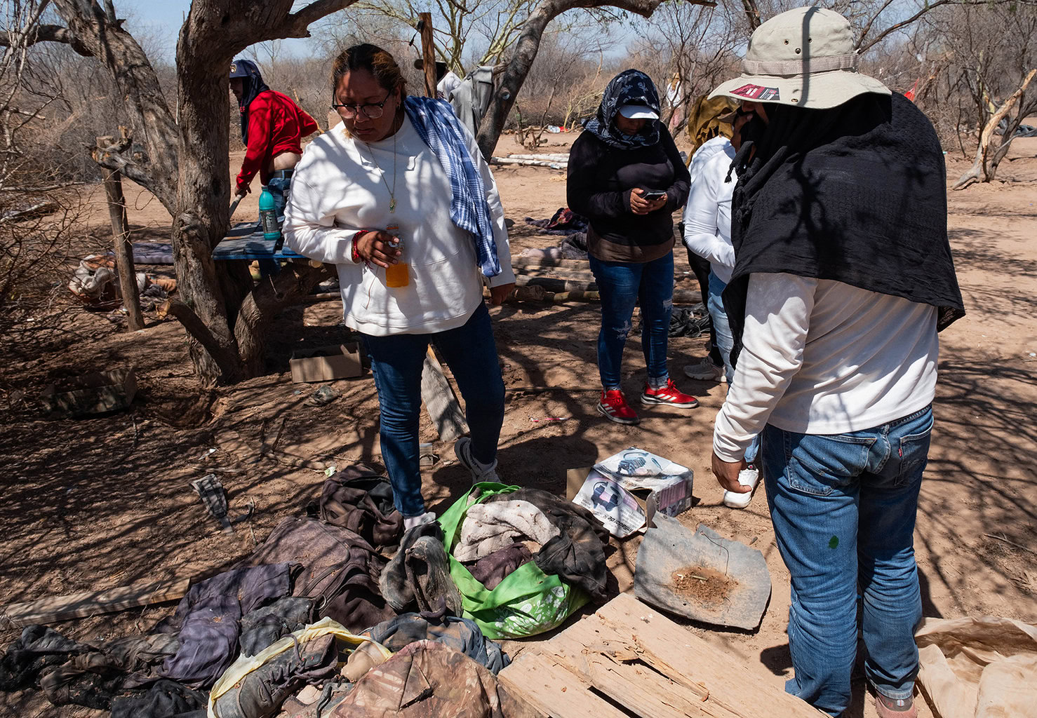 A group of people gather outdoors in a dry, wooded area, surrounded by various items and clothing on the ground. Some are standing, while others appear engaged in an activity.