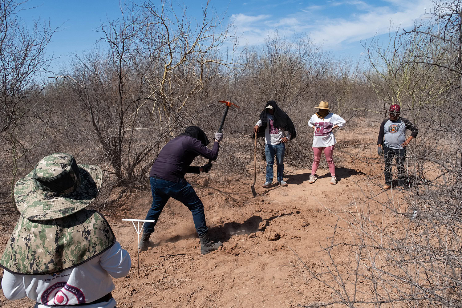 A group of people is digging in a dry, sparse landscape. One person uses a pickaxe while others observe.