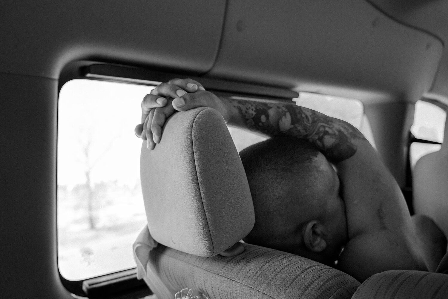 A person with tattooed arms rests their head and arms on a car seat, looking out the window. Black and white photo.