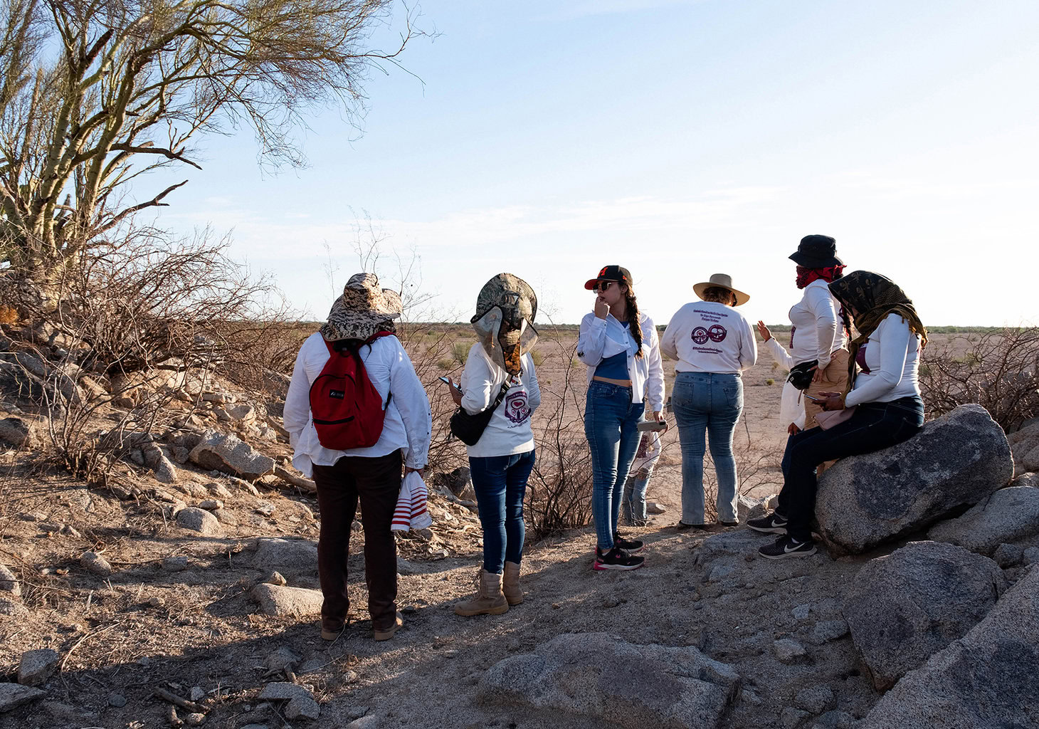 A group of people in white shirts and hats stand on rocky terrain near sparse bushes, looking out at a dry landscape.