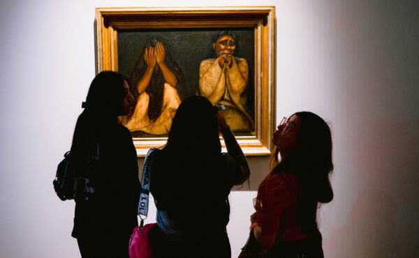 Three people stand in front of a painting depicting two figures, one covering their face and the other with a contemplative expression.