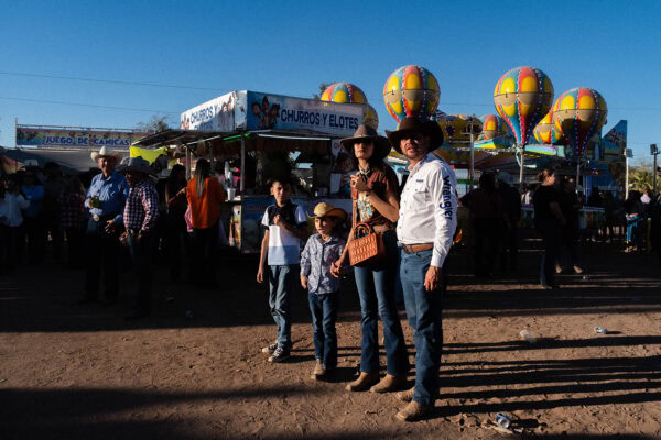A family in western attire stands together at an outdoor fair with colorful balloons and food stalls in the background.