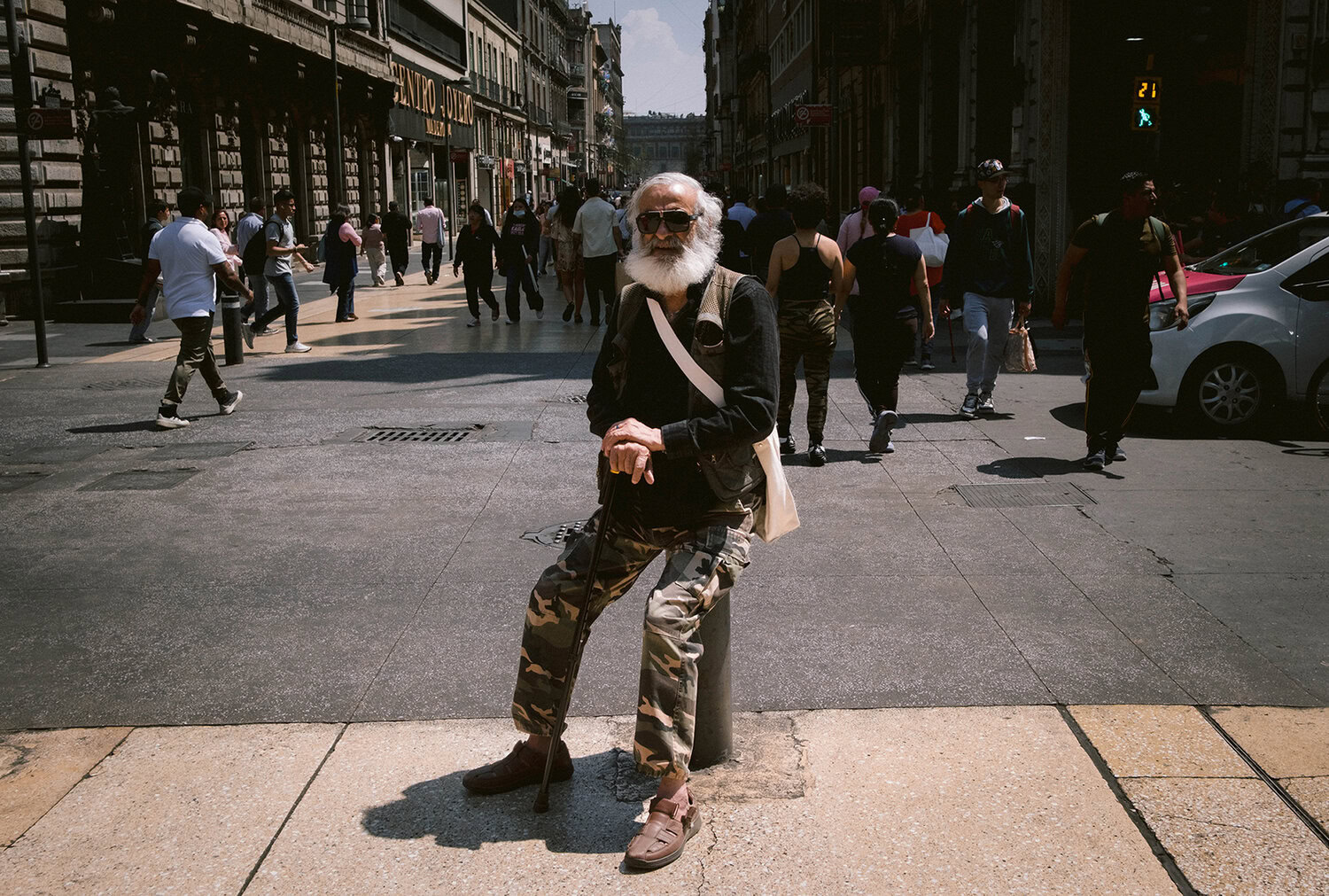 An elderly man with a white beard, wearing sunglasses and camouflage pants, sits on a pole in a city street as people walk by.