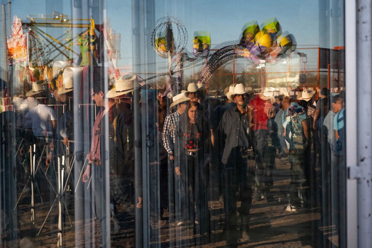 Reflection of people wearing cowboy hats at a carnival, seen through glass with visible rides and balloons in the background.