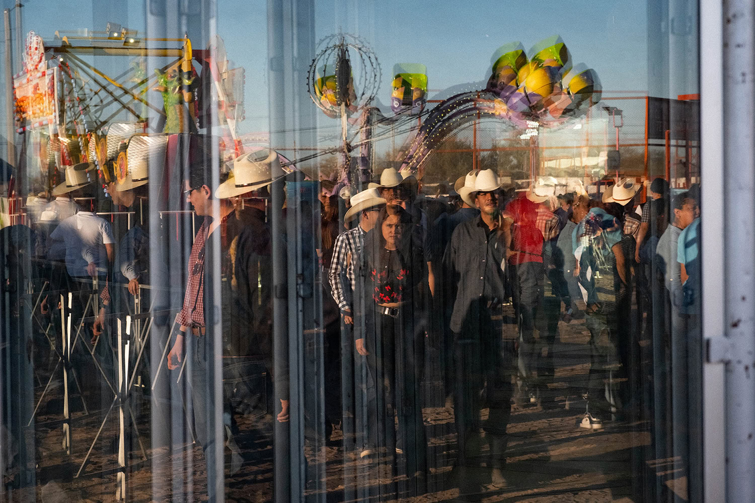 Reflection of people wearing cowboy hats at a carnival, seen through glass with visible rides and balloons in the background.