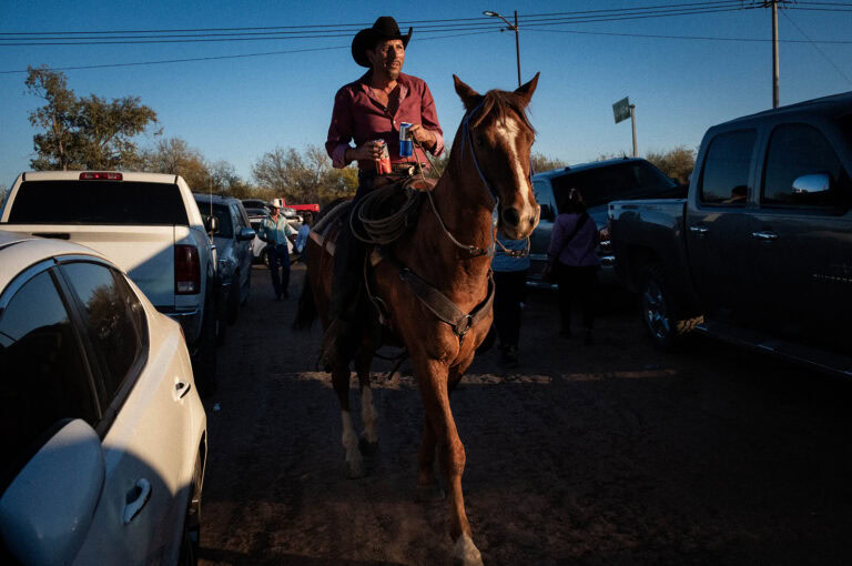 Man in a cowboy hat riding a horse through a parking area with parked cars on either side, holding a can.