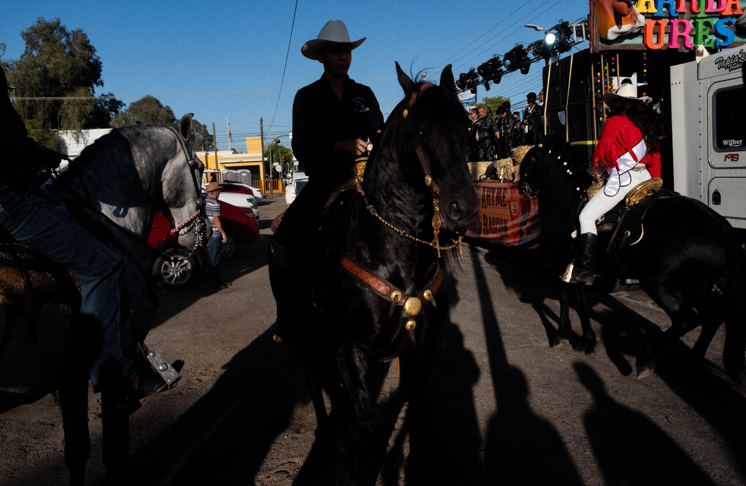 Several people riding horses participate in a street parade during daylight, with a decorated float and spectators visible in the background.