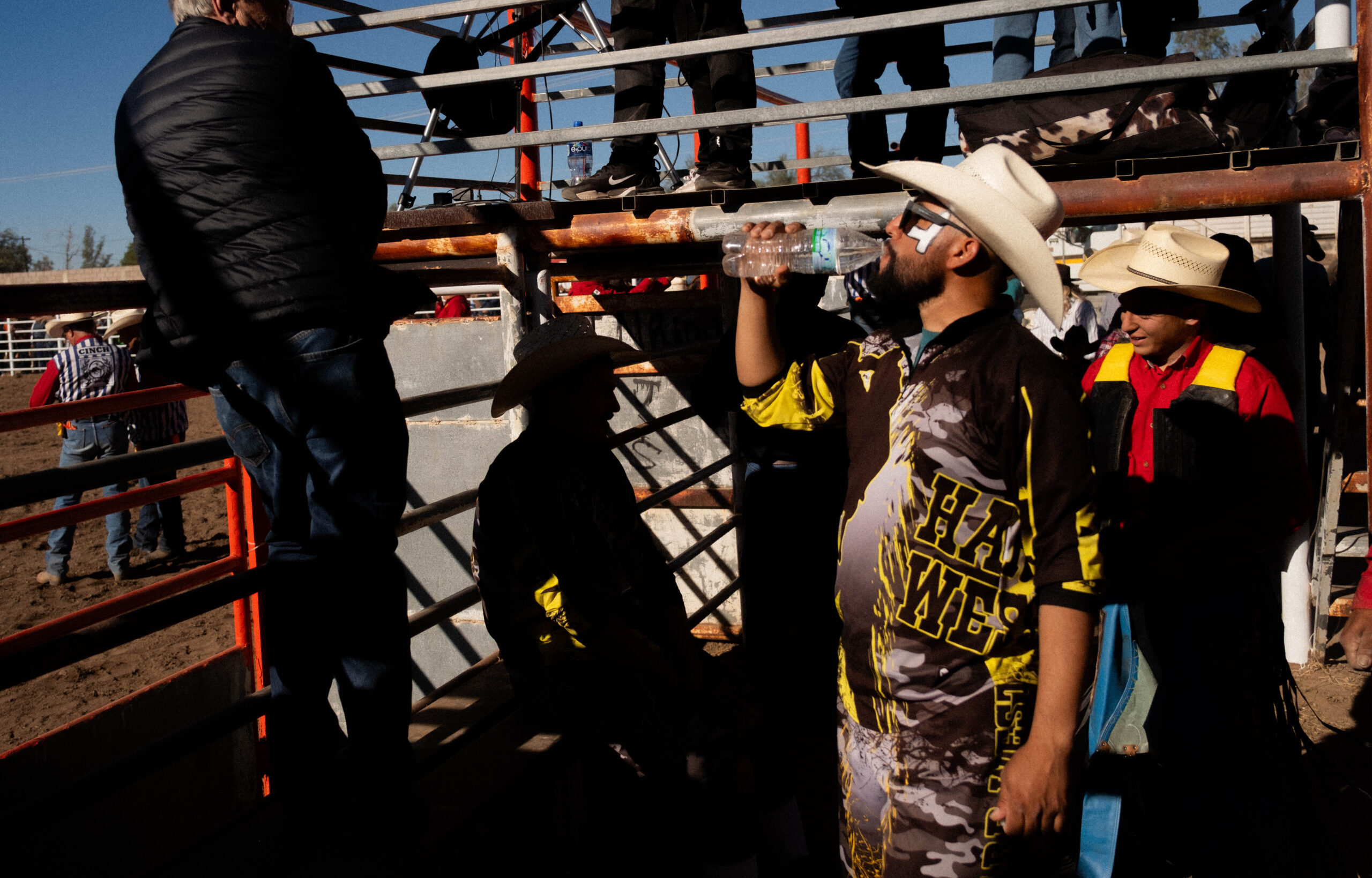 A man in a cowboy hat drinks from a water bottle near a rodeo arena gate, with other people in western attire standing and sitting nearby.
