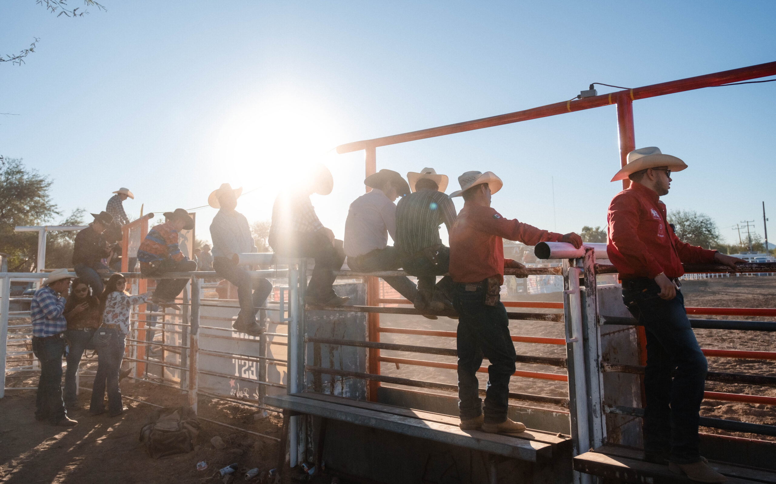 A group of cowboys in hats and boots sit and stand on a rodeo fence, with sunlight shining behind them at an outdoor arena.