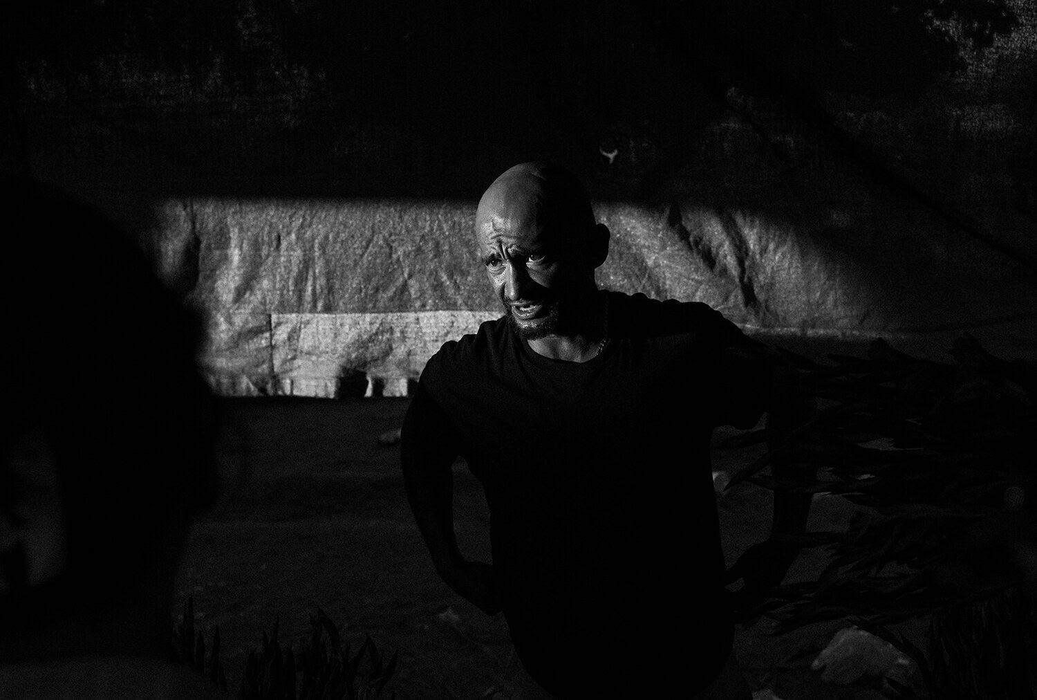 A bald man in a dark shirt stands in dramatic lighting, casting shadows on his face, with another person partially visible in the foreground.