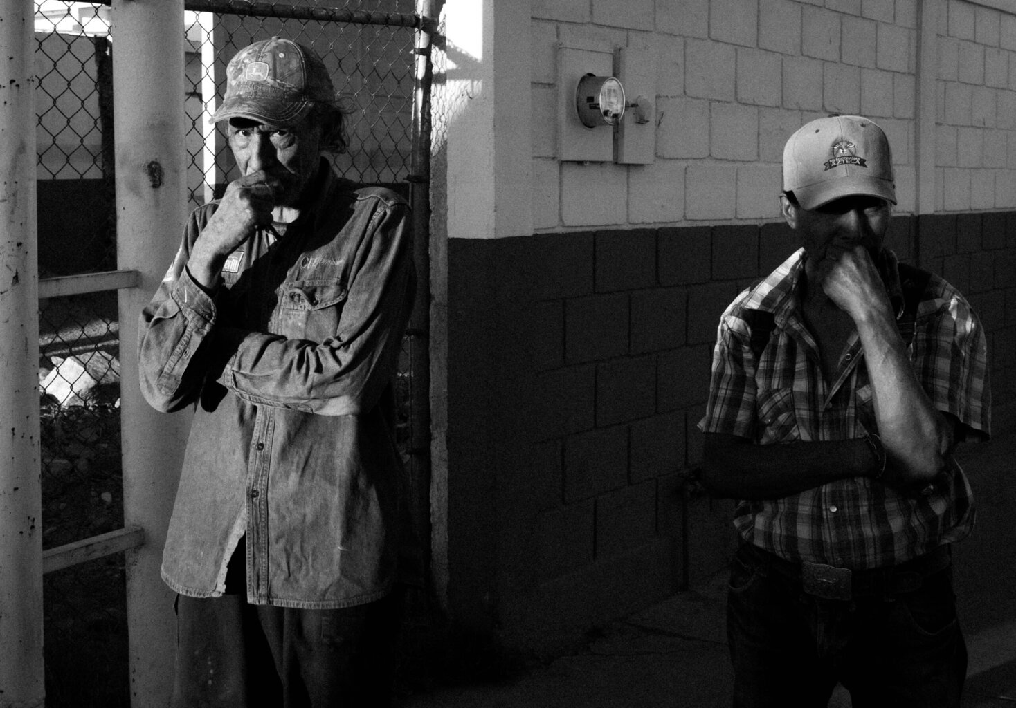 Two men in caps stand outside by a fence and wall, both with one hand to their mouths, wearing casual shirts, in a black-and-white photo.
