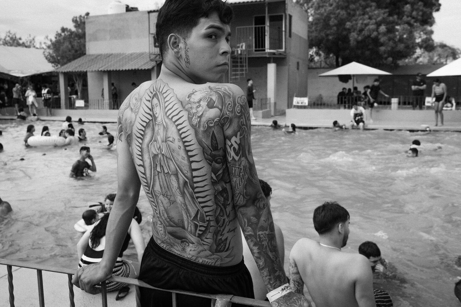 A young man with detailed tattoos on his back stands by a crowded outdoor pool, looking over his shoulder. Other people are swimming and sitting around the pool area.