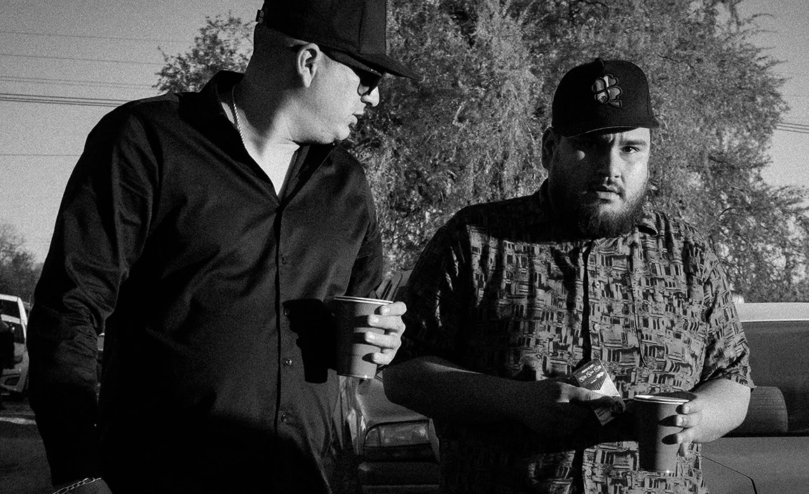 Two men wearing caps stand outdoors holding cups. One man wears sunglasses and a dark shirt while the other has a patterned shirt and looks at the camera. Vehicles and trees are visible in the background.