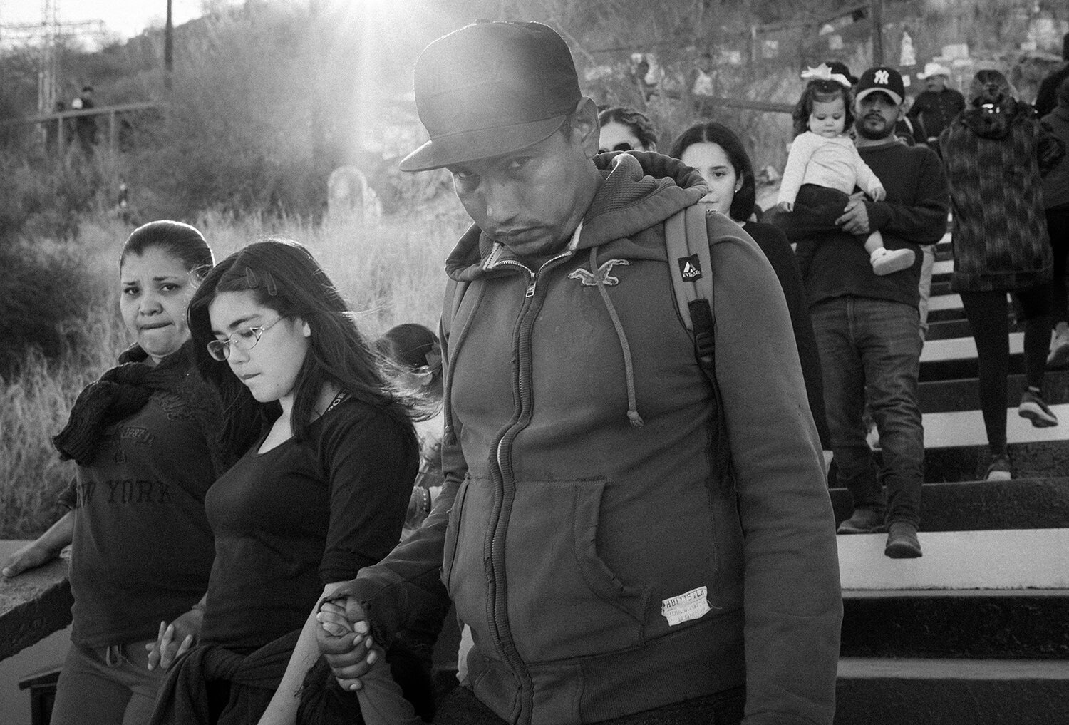 A group of people, including adults and children, walk closely together outdoors on steps, illuminated by sunlight from behind.