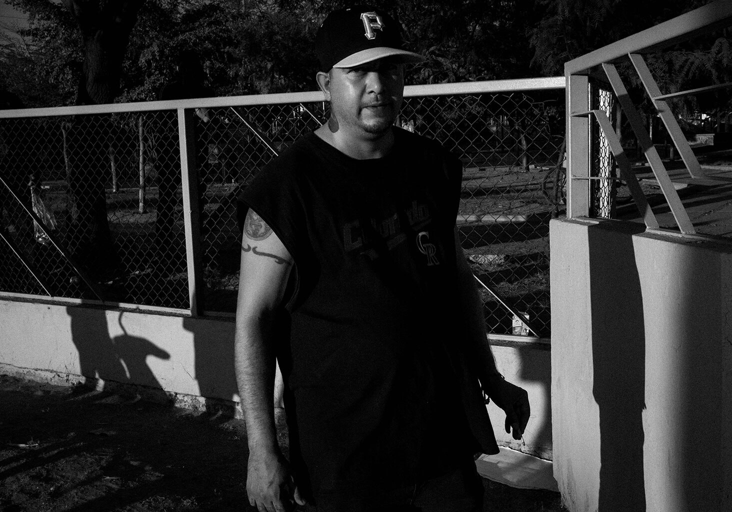 A man in a sleeveless shirt and baseball cap stands near a fence and gate, with sunlight casting shadows on the wall behind him.