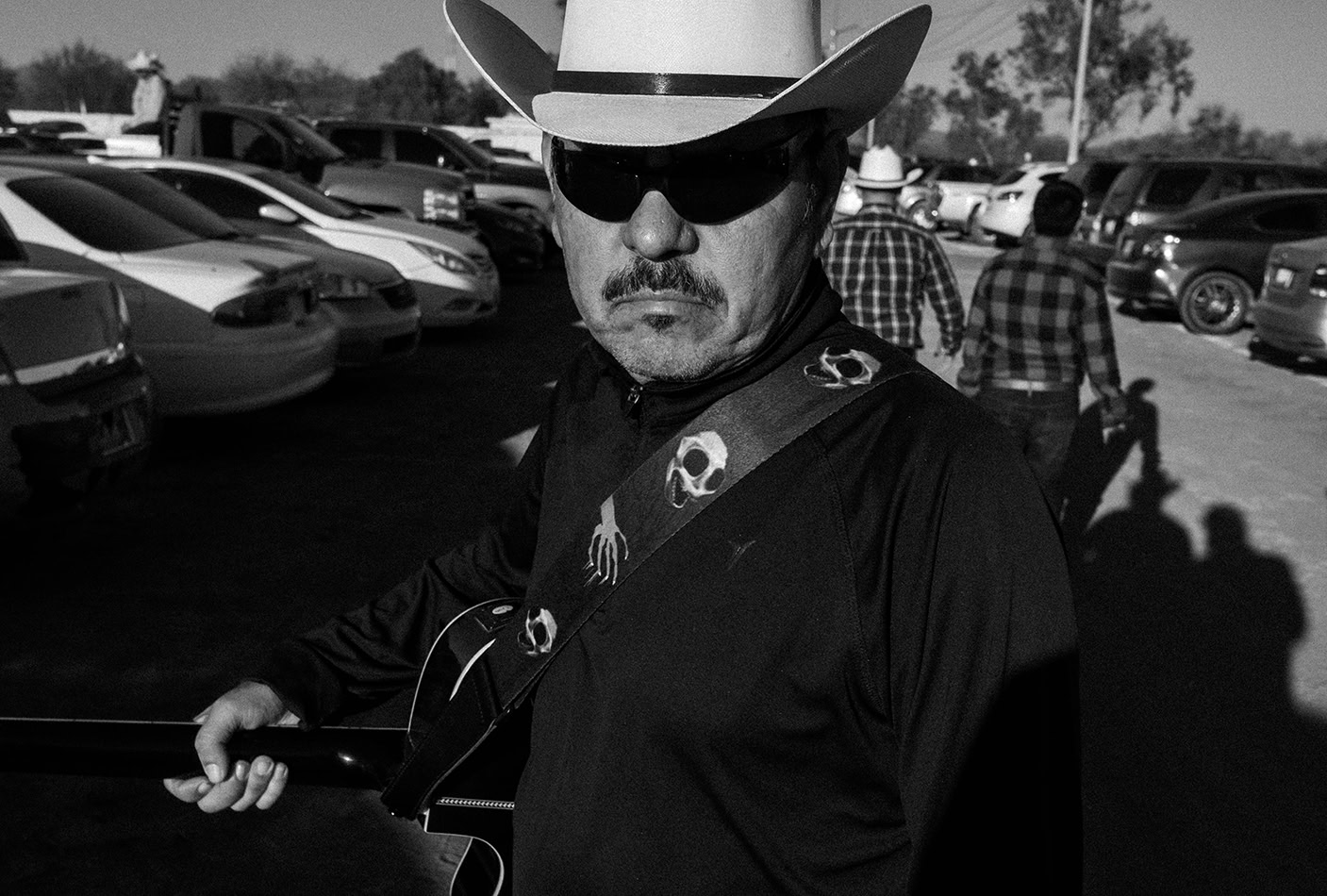 Man wearing a cowboy hat and sunglasses holds a guitar in a parking lot, with several cars and people in the background.