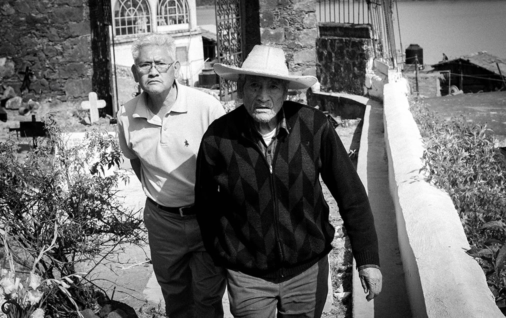 Two older men walk outdoors along a narrow path near a stone building, with one man in a hat leading and the other following closely behind.
