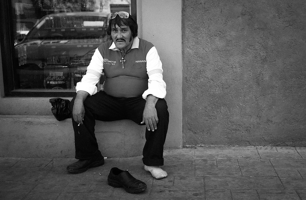 A man sits on a sidewalk with one shoe off and bare foot, beside a wall and window, looking at the camera.