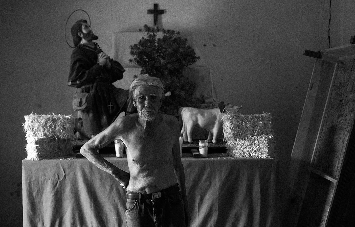 An older shirtless man stands in front of a rustic altar with religious statues, hay bales, candles, and a small cross.
