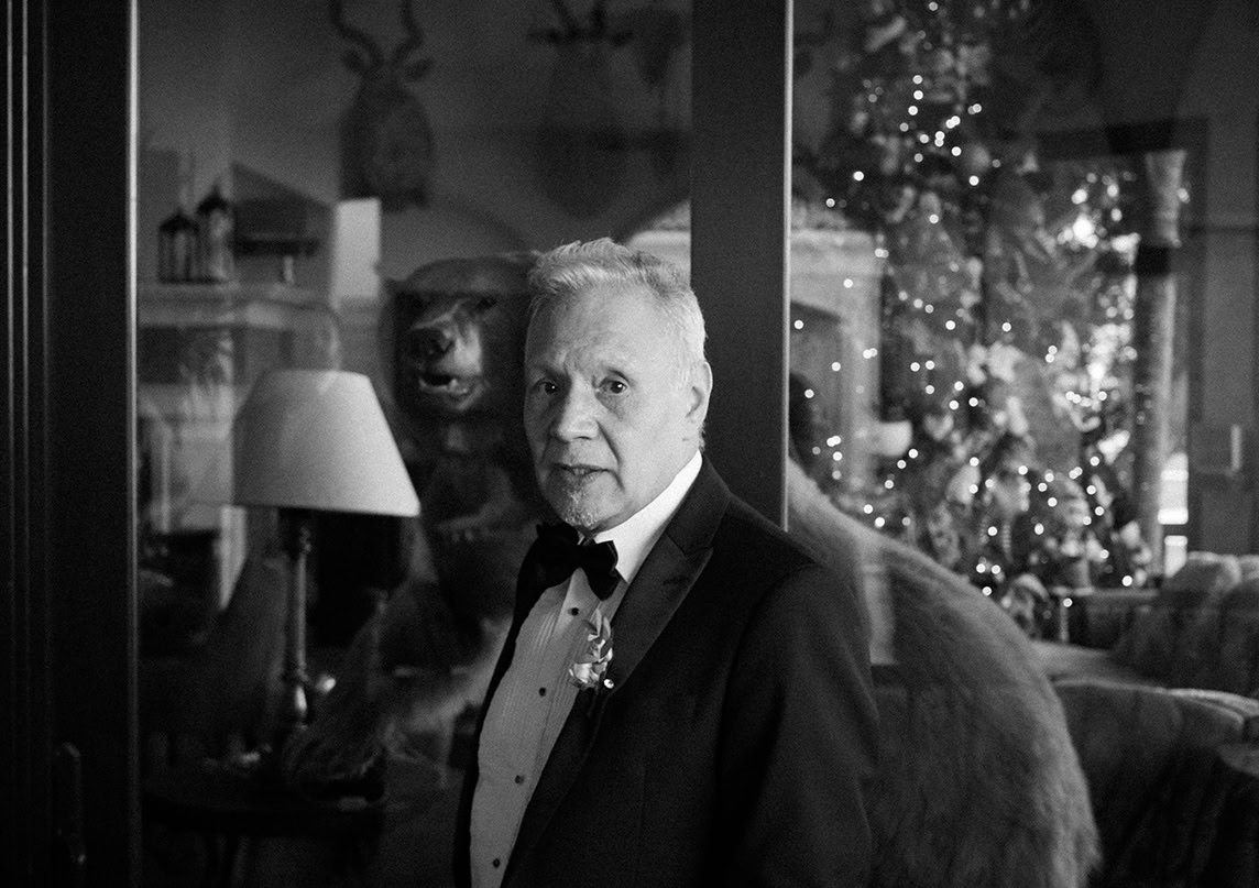 An older man in a tuxedo stands indoors near a glass wall, with a decorated Christmas tree and a bear taxidermy in the background.