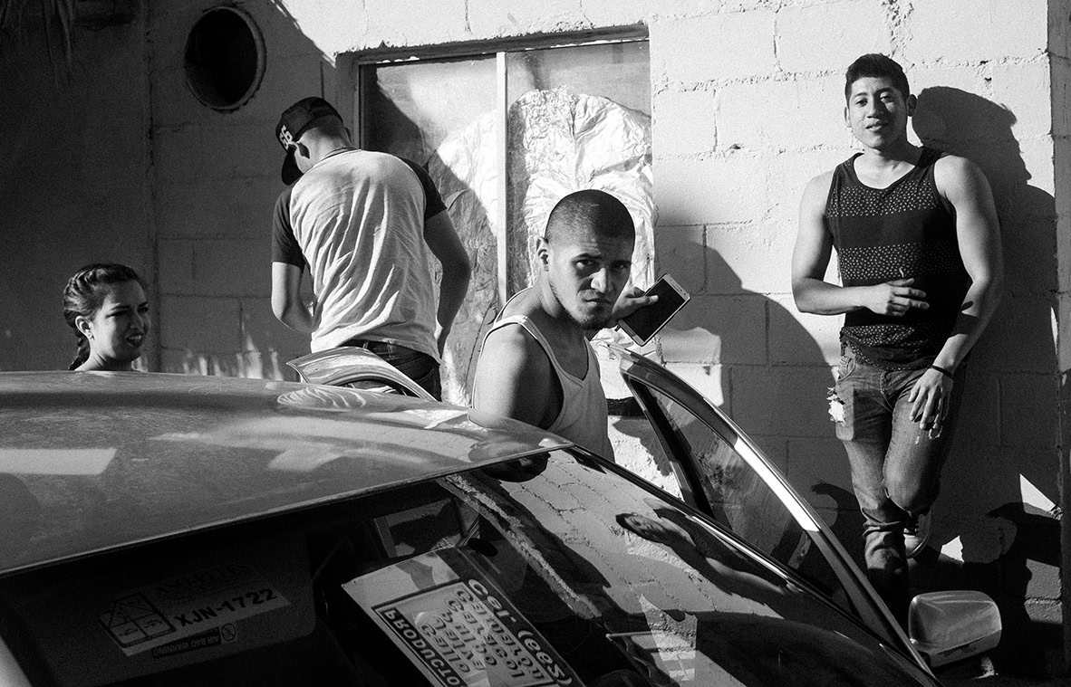 Four young adults gather near a car and a building; one stands by the car door, others lean against the wall, and sunlight casts sharp shadows.