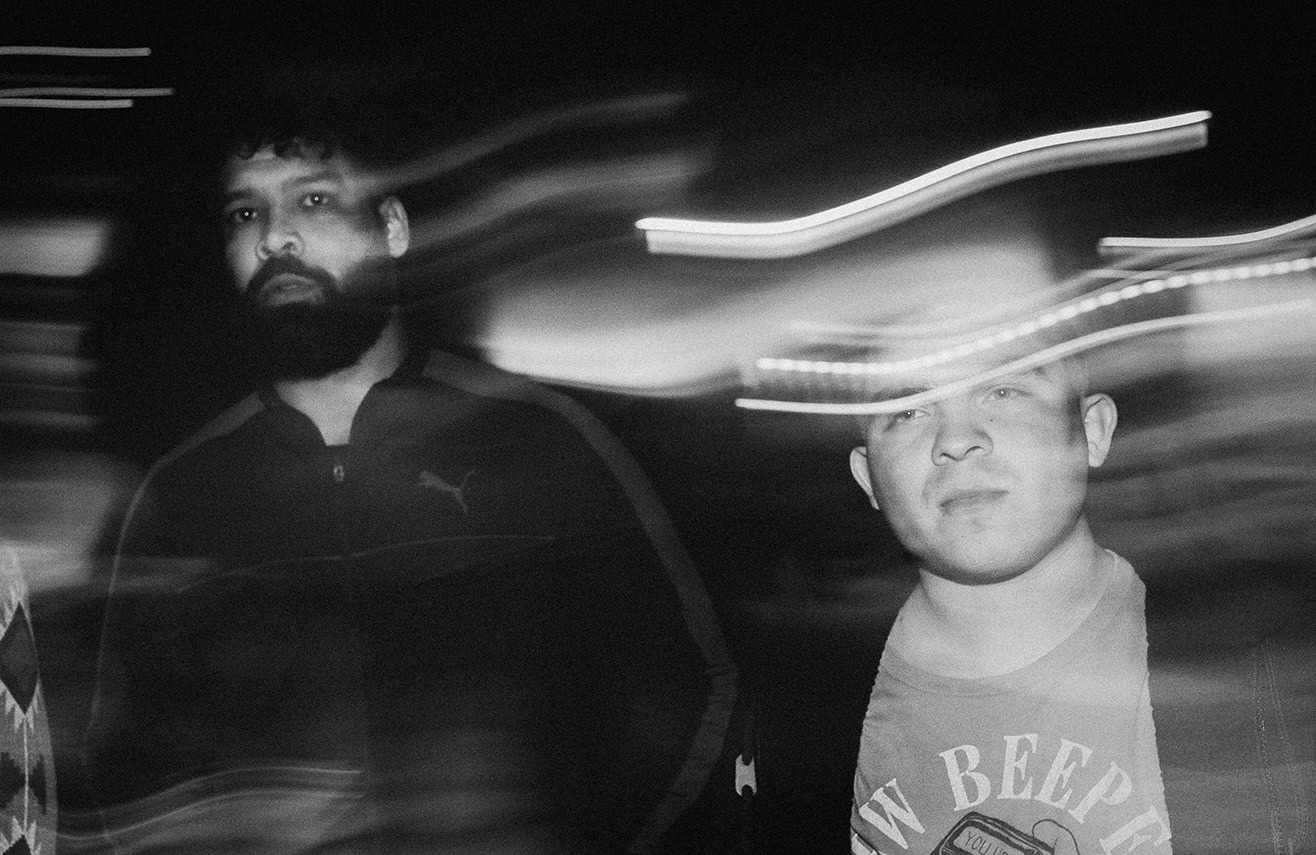 Two men stand in a dim setting with light streaks creating motion blur across the black-and-white photo.