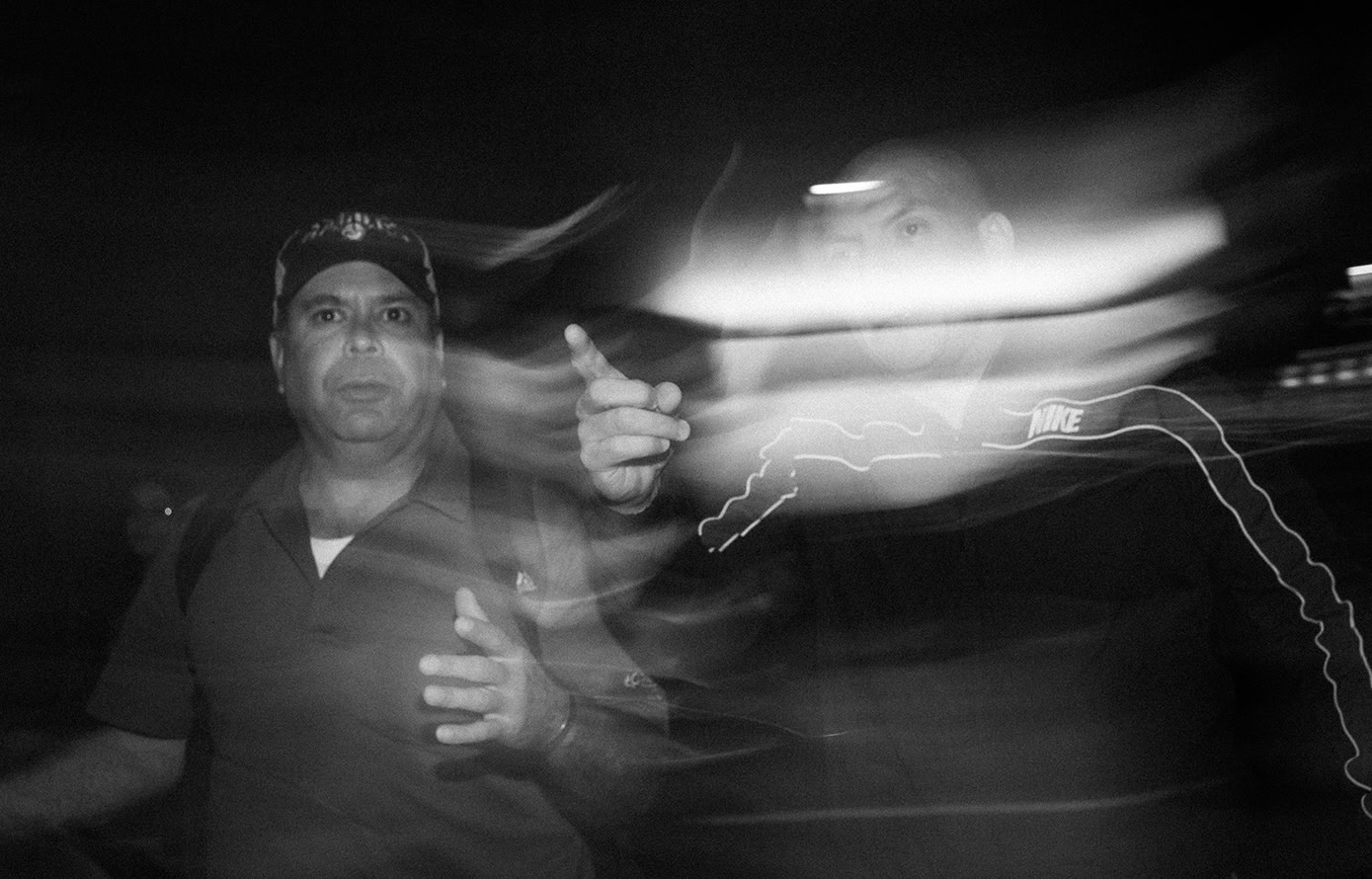 Two men stand at night; one gestures toward the camera. Motion blur and light streaks partially obscure the scene, creating a sense of movement and urgency.