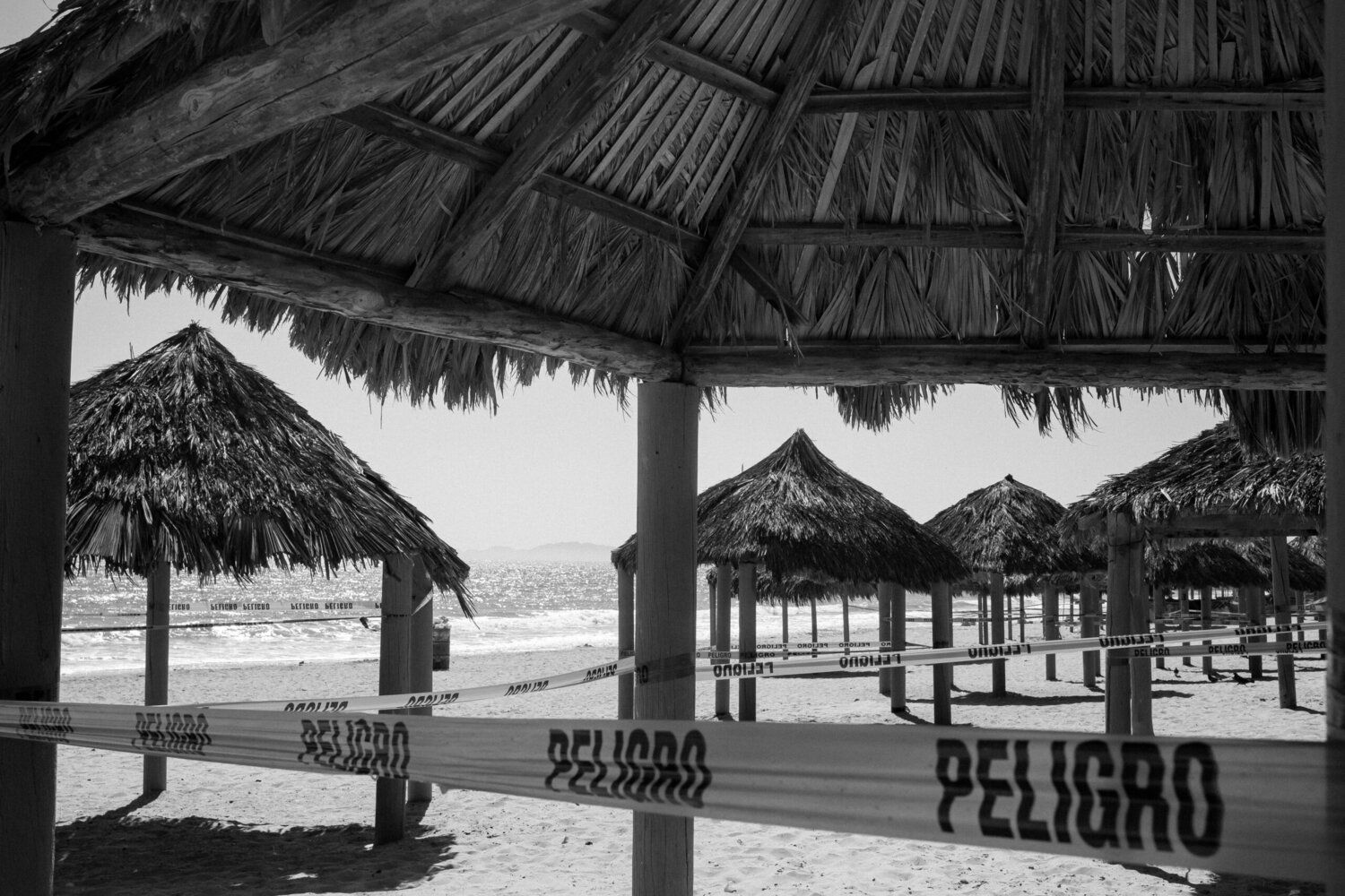 Thatched beach umbrellas are closed off with caution tape labeled “Peligro” on a sandy beach, with the ocean and mountains visible in the background.
